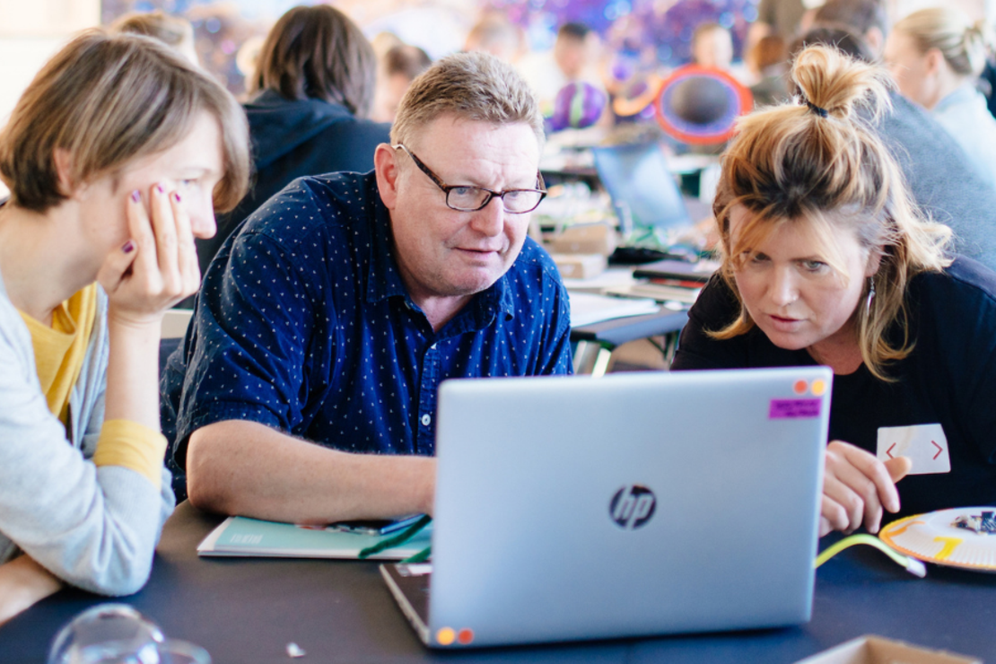 Drei Personen schauen auf einen Laptop. Vor ihnen liegen technische Bauteile.