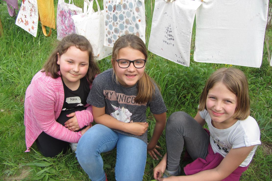 Drei Kinder hocken im Gras vor mit Pflanzenfarbe bemalten Stofftaschen