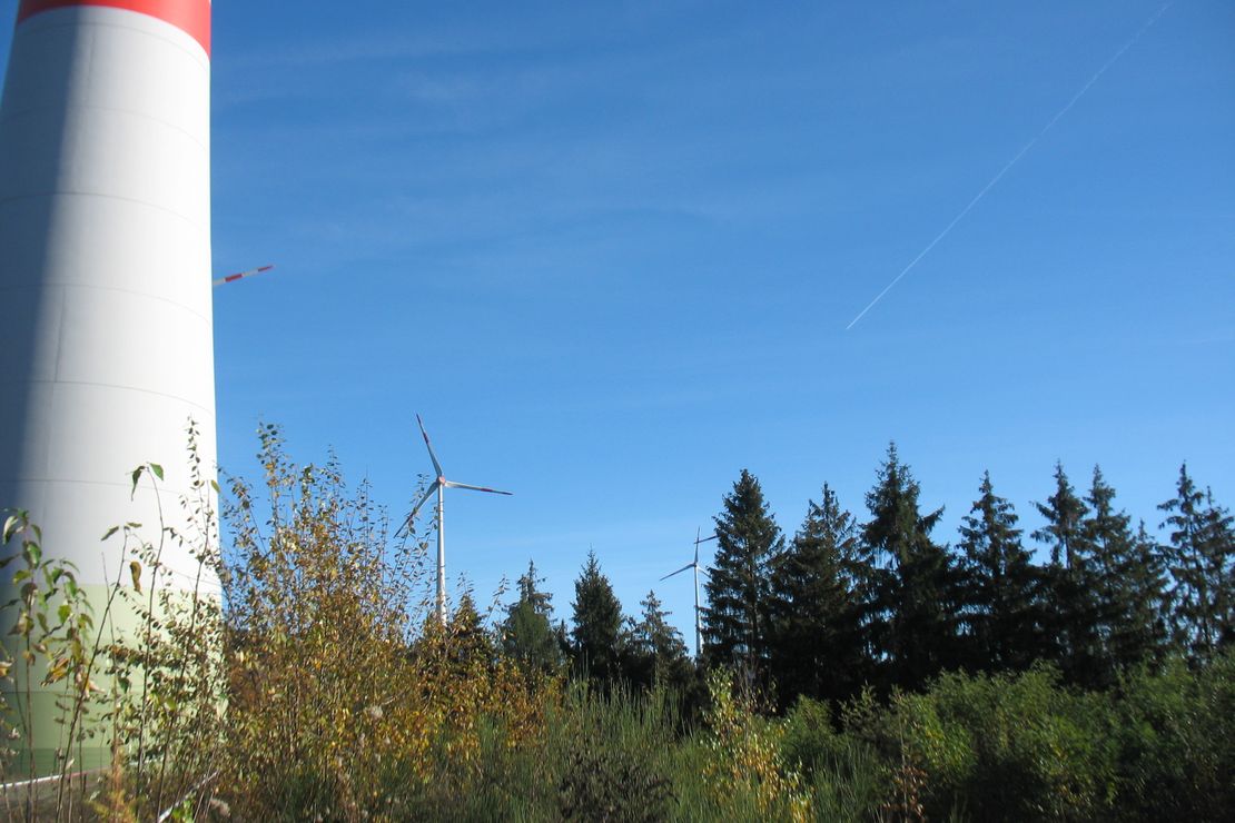 Vor einem Fichtenbestand sieht man neben dem Sockel einer Windkraftanlage die Rotorblätter weiterer Windkraftanlagen über den Baumspitzen