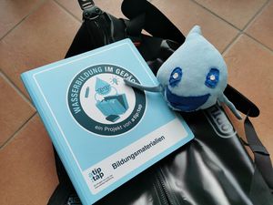 Der Bildungsordner liegt neben dem Maskottchen des Bildungsprogramms, ein knuddeliges Regentropfenwesen