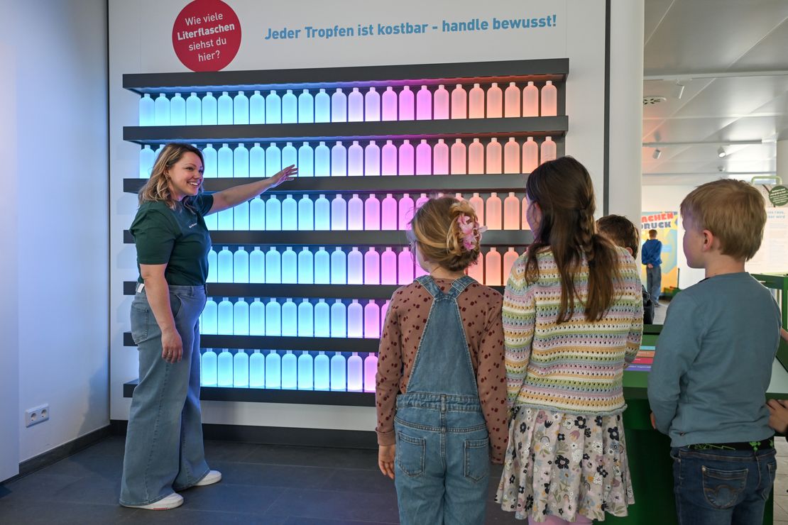 Ein Mensch steht vor einem Expomat der WasserWerkstatt und erklärt dieses den Besucherinnen.