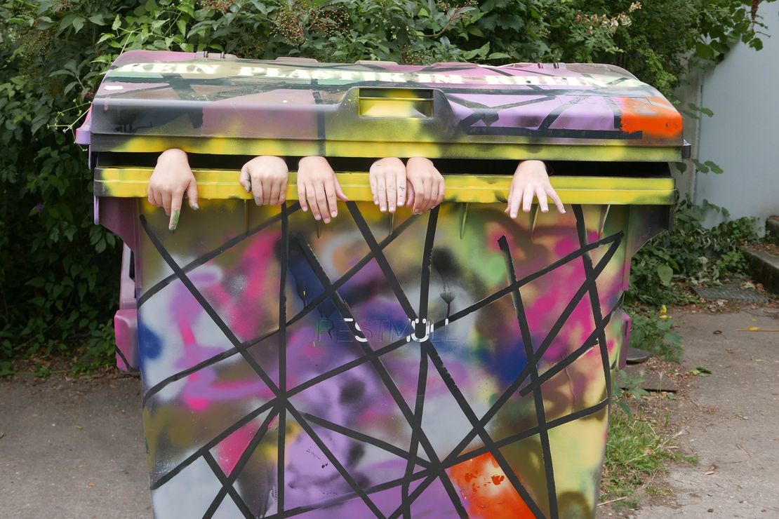 zu sehen ist ein Müll-Container, der mit bunten Grafittis verziert wurde. Oben ist der Schriftzug "Kein Plastik im Rhein" zu lesen. Aus dem Container  schauen Kinderhände .