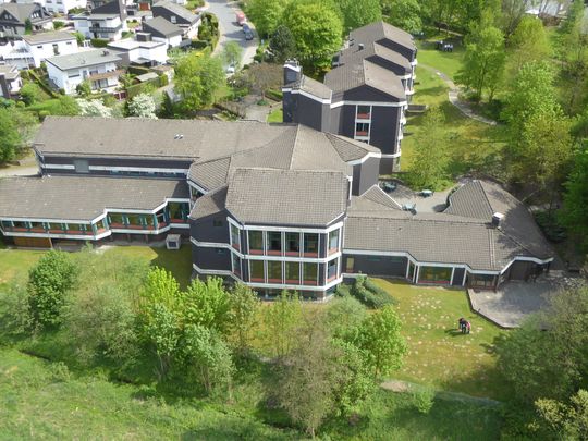 Auf dem Bild ist das Tagungshaus der Akademie Biggesee aus der Luft zu sehen. Man sieht ein mehrstöckiges Gebäude mit mehreren Gebäudeteilen umgeben von einer grünen Gartenlandschaft.7