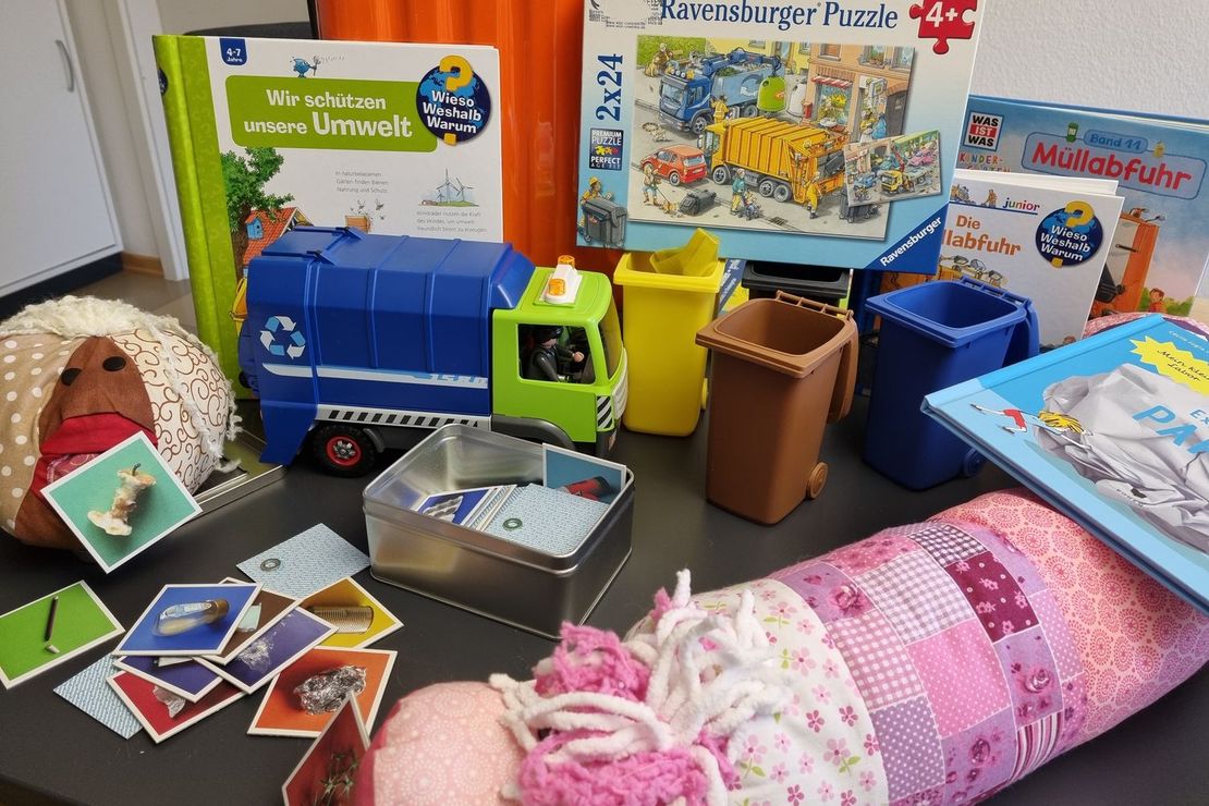 Das Foto zeigt die Arbeitsmaterialien, die der Koffer enthält: Kuschel-Kompostwurm Lombi® und das Methan-Baktinchen®, mit den zugehörigen Magnetkärtchen, kleine Mülltonnen, ein Müllfahrzeug und Bücher