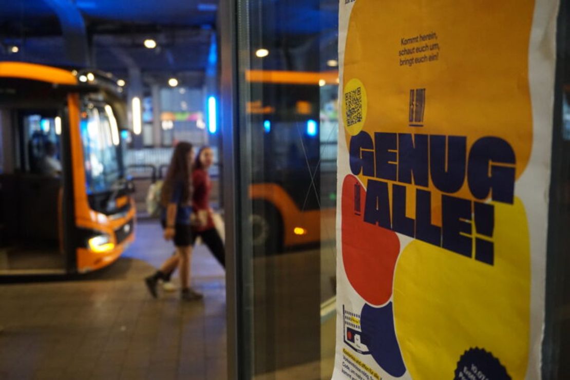 Ein Plakat mit dem Text "Es gibt genug für alle" hängt im Fenster, dahinter laufen Menschen und es ist ein Bus zu sehen.