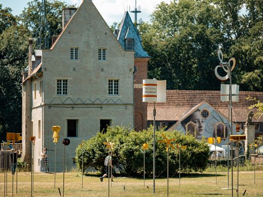 Von Schüler:innen erstellte kleine Windrädern, die im Schlosspark als Kunstwerke aufgestellt wurden, sind vor dem Schlossensemble des Schlosses Senden zu sehen.