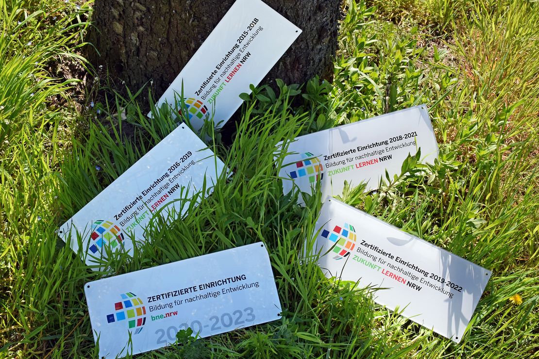Fünf Auszeichnungsschilder der BNE-Zertifizierung NRW sind auf einer Wiese an einen Baumstamm gelehnt.