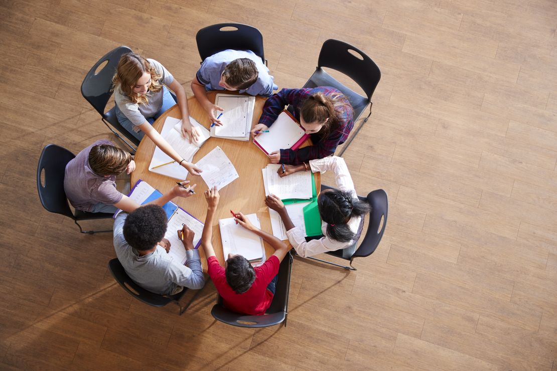 Aus der Vogelperspektive: sieben junge Menschen sitzen an einem runden Tisch, arbeiten zusammen und tauschen ihre Papiere und Ideen aus.