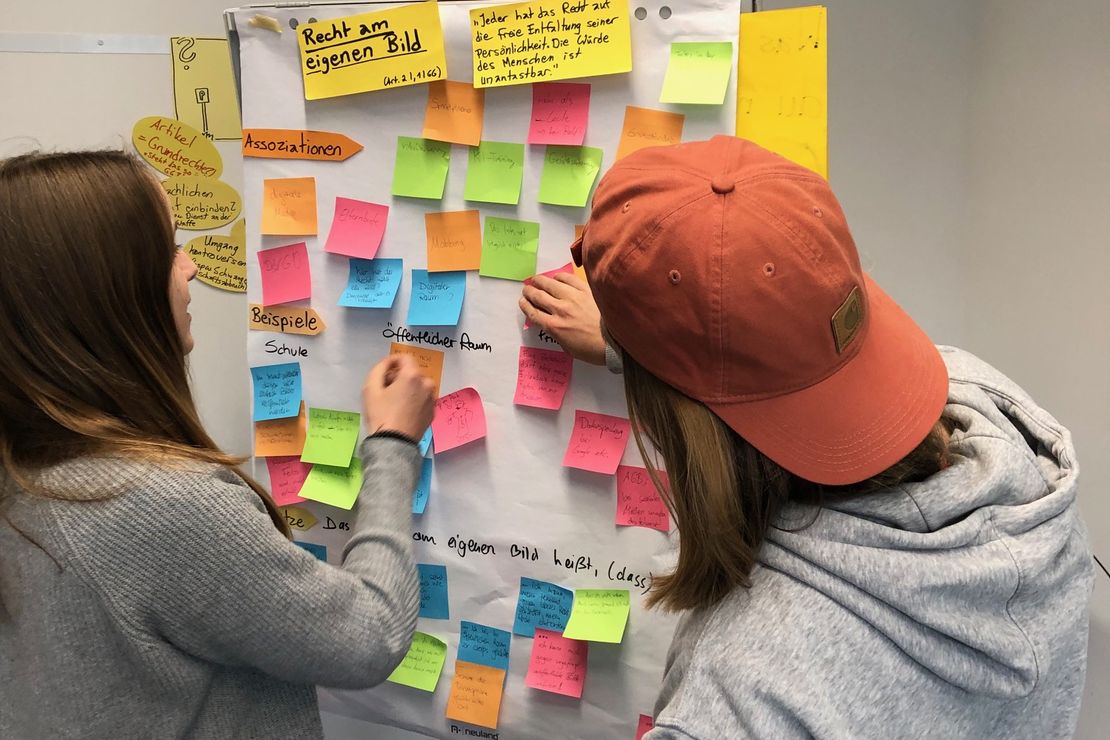 Auf dem Bild sind zwei, an einem GrundgeSÄTZE-Workshop teilnehmende Schüler*innen abgebildet, die Post-its beschriften und an einem Flipchart-Papier sortieren.