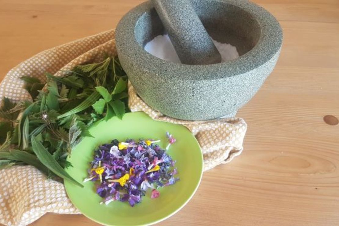 frische Kräuter und bunte Blüten liegen vor einem mit Salz gefüllten Mörser