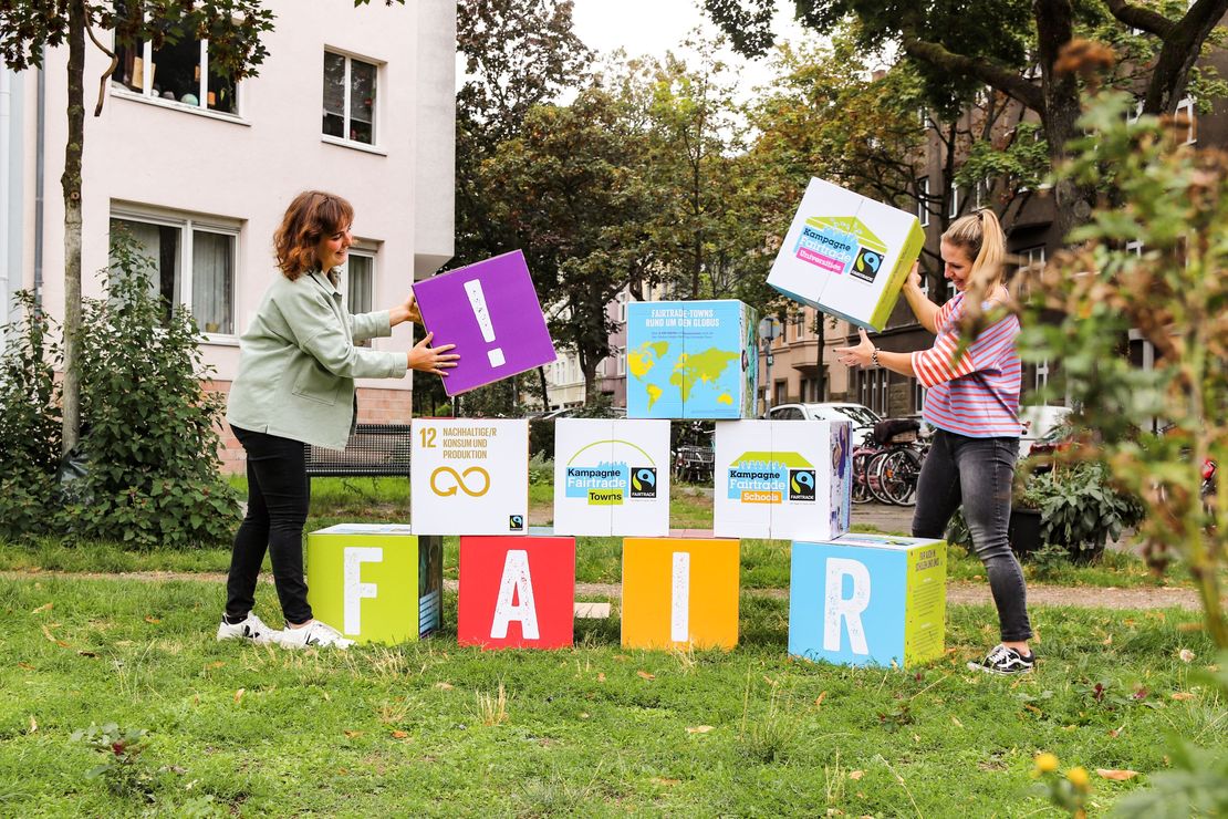Zwei Frauen stapeln Pappwürfel. Darauf zu lesen ist "Fair", das SDG 12 und die Logos der Kampagnen Fairtrade-Town, Fairtrade Schools und Fairtrade Universities.