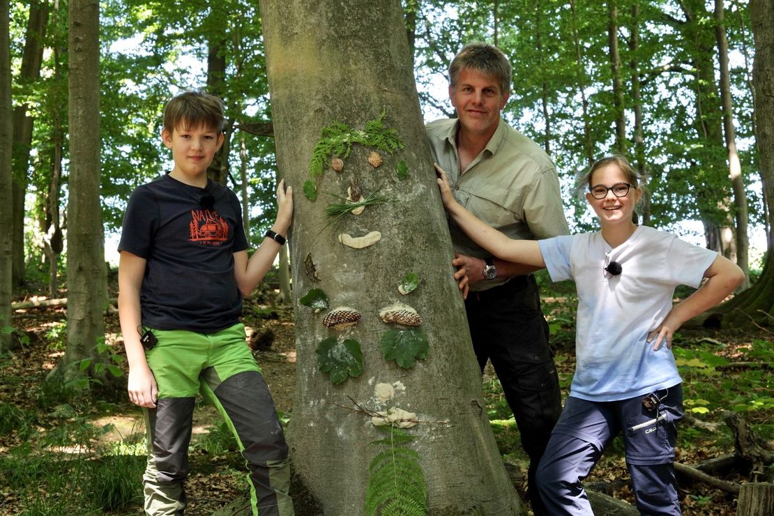 Förster Stefan Nolte und die Kinder Klara und Simon schauen hinter einem Buchenstamm hervor, auf dem sich ein selbstgemachtes Gesicht aus Ton befindet.