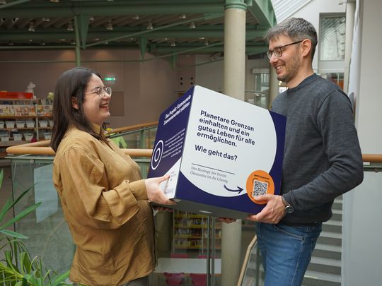 Zwei Personen stehen in einer Bibliothek und lachen sich an. Gemeinsam halten sie einen großen Würfel in der Hand, auf dem Infos zu einem Projekt für kommunale Nachhaltigkeit stehen.