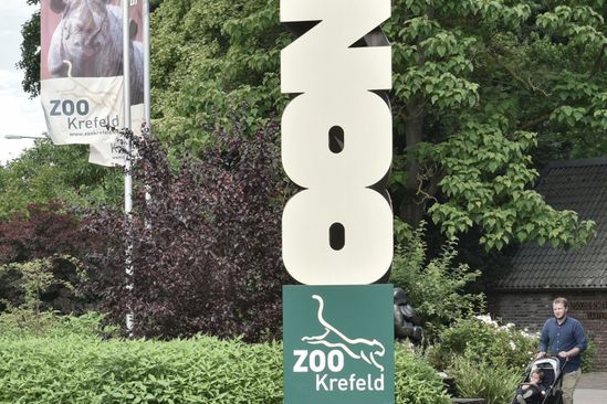 Der Eingangsbereich des Zoo Krefeld mit Fahnen und einem Besucher mit Kinderwagen ist zu sehen.