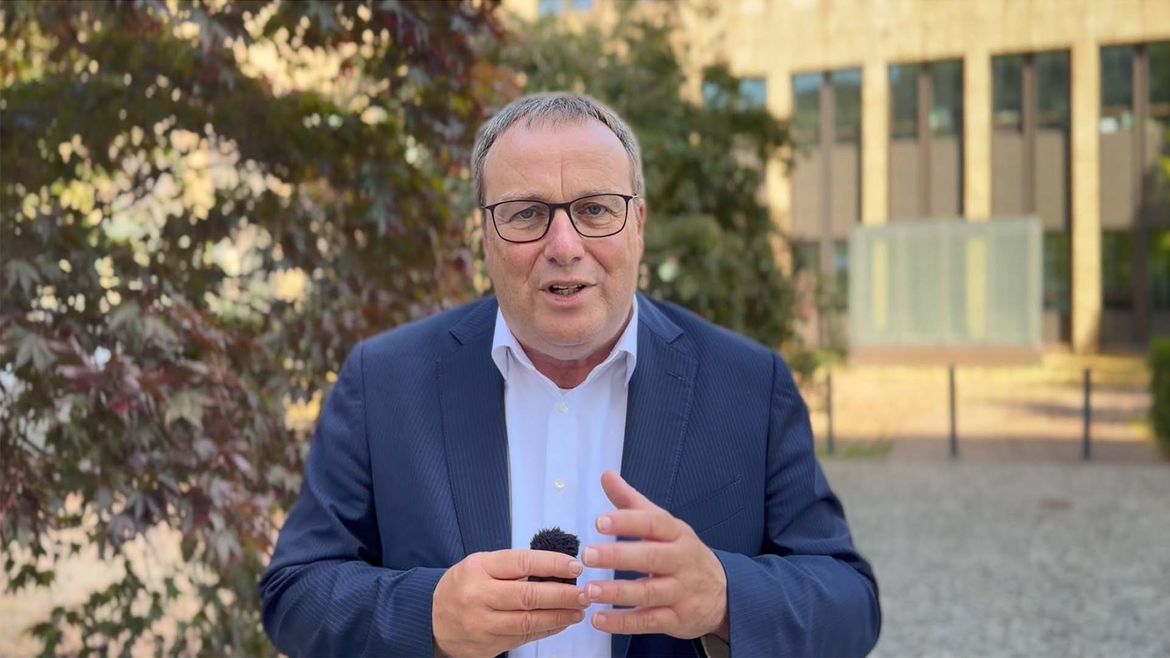 Herr Minister Oliver Krischer steht draußen vor dem Gebäude des Minsteriums und blickt in die Kamera. Er trägt ein blaues Sakko. Im Hintergrund ein Strauch.