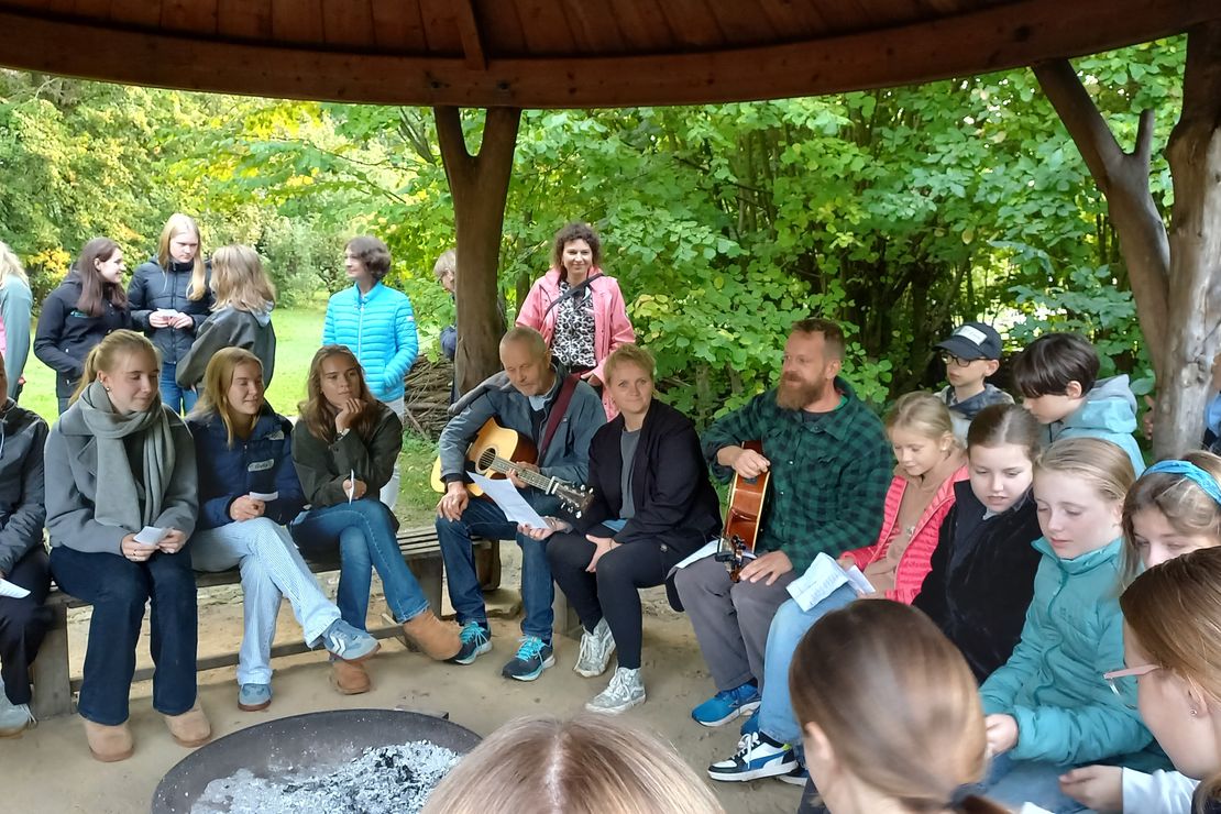 Lehrkräfte und Schüler*innen sitzen an einer überdachten Feuerstelle und singen gemeinsam mit Gitarrenbegleitung ein Lied