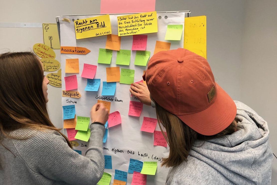Auf dem Bild sind zwei, an einem GrundgeSÄTZE-Workshop teilnehmende Schüler*innen abgebildet, die Post-its beschriften und an einem Flipchart-Papier sortieren.