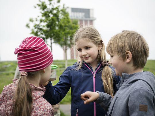 Drei Kinder stehen zusammen. Ein Kind hält eine Becherlupe hoch, auf die alle schauen. Im Hintergrund ist ein Hügel, ein Baum und ein Förderturm zu sehen.