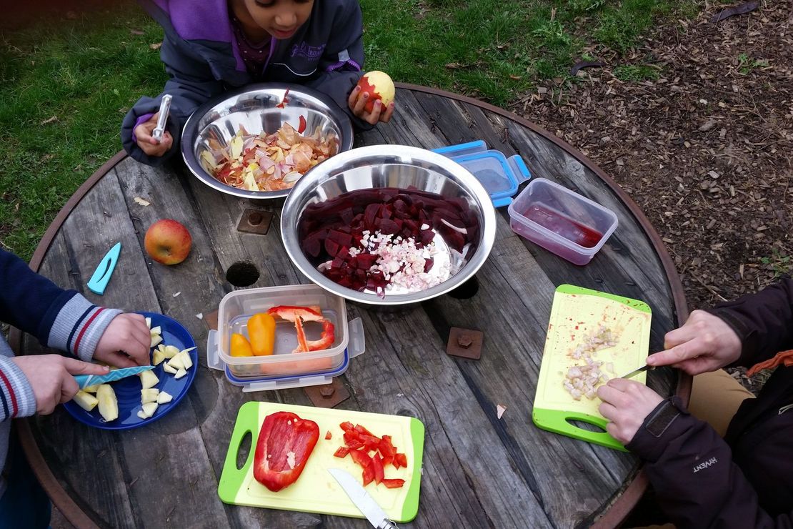 Zwei Kinder und eine Erwachsene schneiden draußen an einem Holztisch Gemüse für einen leckeren Salat