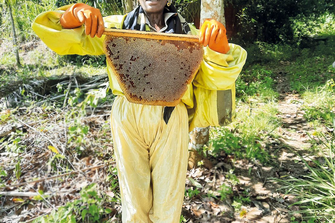 Eine Frau aus Kamerun im Imkerschutzanzug hält eine Bienenwabe in die Kamera