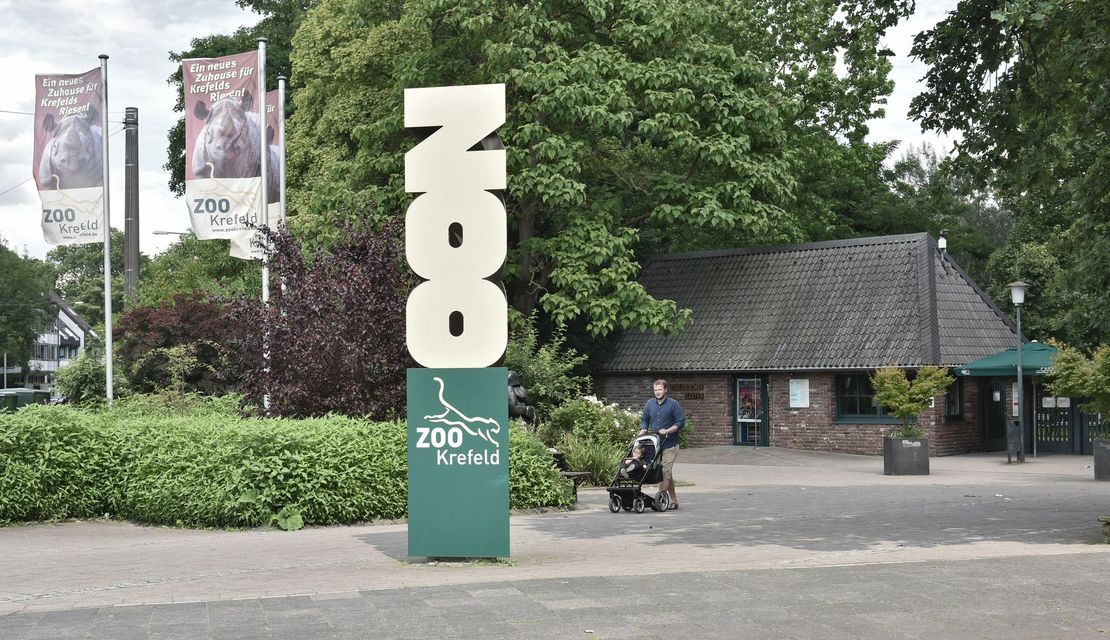 Zu sehen ist der Eingangsbereich des Zoo Krefeld mit Besuchern, Kassenhäuschen und Fahnen.