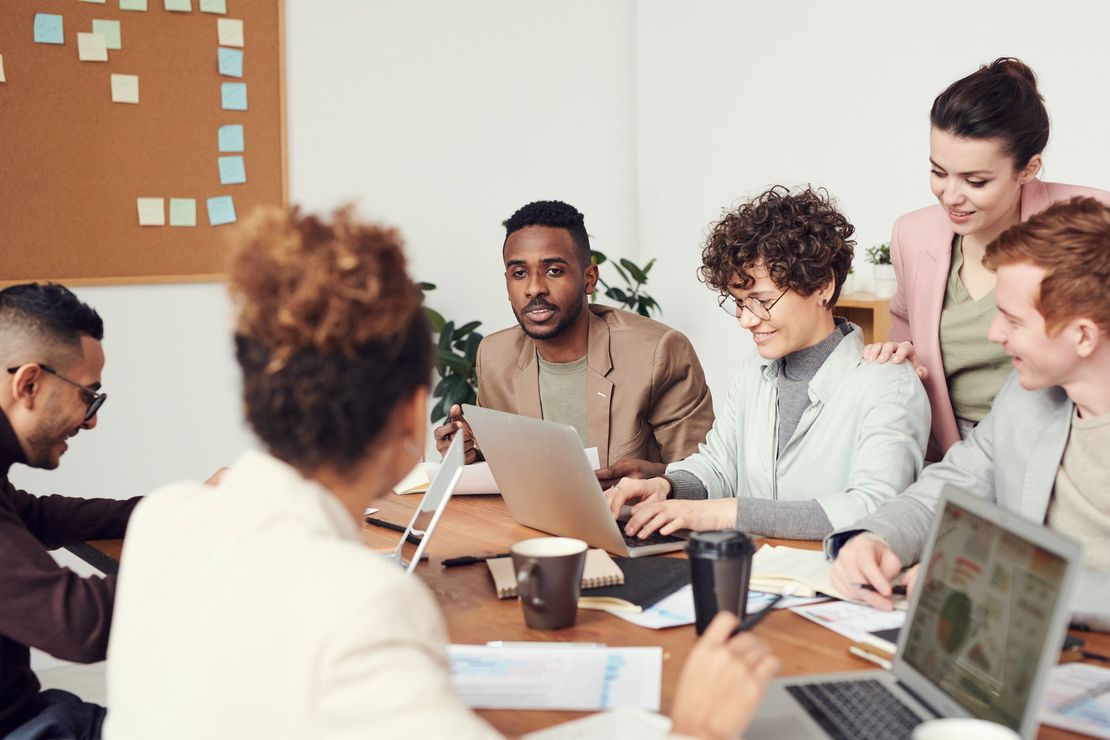 Eine Gruppe von Menschen sitzt mit Notizzetteln, Notebooks und Kaffeetassen zusammen an einem Tisch und arbeitet zusammen. An einer Tafel an der Wand hängen verschiedene Haftnotizen.