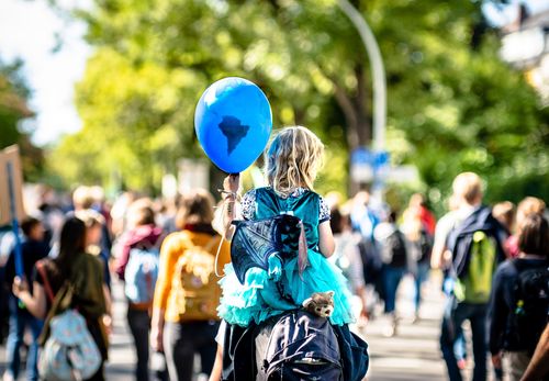 Ein Schriftzug "Global - lokal. BNE 2030 in der praktischen Bildungsarbeit" ist halbtransparent über ein Foto gelegt. Darauf ein Kind mit Luftballon, Huckepack auf den Schultern eines Erwachsenen. Der Luftballon ist blau und einer Weltkugel nachempfunden. Das Kind trägt ein Feen-Kostüm mit Kleid und Flügeln. Der Blick zeigt beide von ihrer Rückseite. Im Hintergrund sind andere Menschen auf einer friedlichen Demonstration zu erkennen. Das Kind hat schulterlanges, blondes Haar und eine weiße Hautfarbe. Es ist ein sonniger Tag.