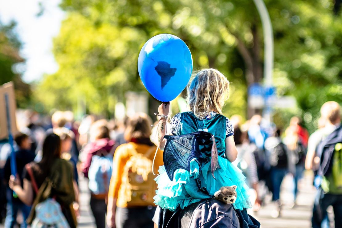 Ein Schriftzug "Global - lokal. BNE 2030 in der praktischen Bildungsarbeit" ist halbtransparent über ein Foto gelegt. Darauf ein Kind mit Luftballon, Huckepack auf den Schultern eines Erwachsenen. Der Luftballon ist blau und einer Weltkugel nachempfunden. Das Kind trägt ein Feen-Kostüm mit Kleid und Flügeln. Der Blick zeigt beide von ihrer Rückseite. Im Hintergrund sind andere Menschen auf einer friedlichen Demonstration zu erkennen. Das Kind hat schulterlanges, blondes Haar und eine weiße Hautfarbe. Es ist ein sonniger Tag.