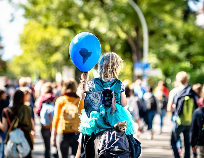 Ein Schriftzug "Global - lokal. BNE 2030 in der praktischen Bildungsarbeit" ist halbtransparent über ein Foto gelegt. Darauf ein Kind mit Luftballon, Huckepack auf den Schultern eines Erwachsenen. Der Luftballon ist blau und einer Weltkugel nachempfunden. Das Kind trägt ein Feen-Kostüm mit Kleid und Flügeln. Der Blick zeigt beide von ihrer Rückseite. Im Hintergrund sind andere Menschen auf einer friedlichen Demonstration zu erkennen. Das Kind hat schulterlanges, blondes Haar und eine weiße Hautfarbe. Es ist ein sonniger Tag.