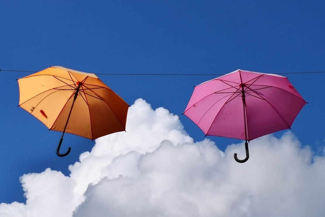 Zwei farbige Regenschirme hängen aufgespannt an einer Leine. Im Hintergrund sind weiße Wolken vor blauem Himmel zu sehen.