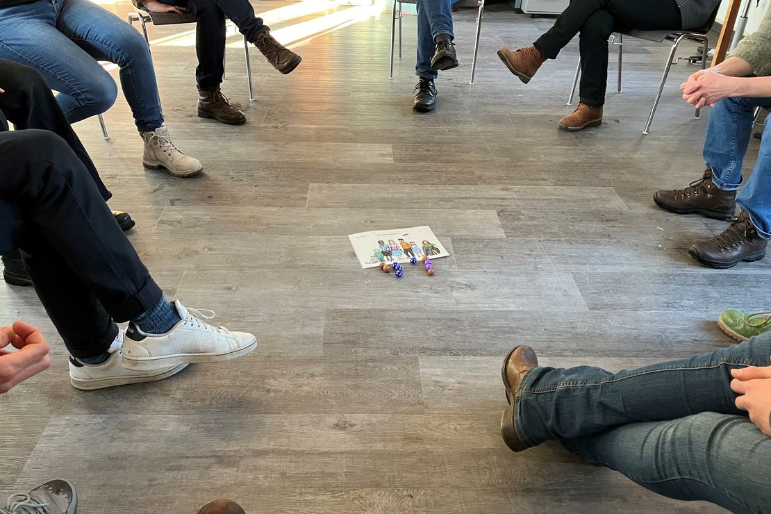 Personen sitzen im Kreis um einer Abbildung herum, welche auf dem Boden liegt und sieben Kinder unterschiedlichen Alters und Aussehens zeigt.