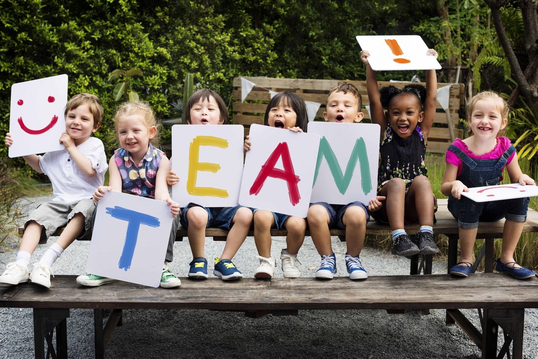 Eine Gruppe von Kindern sitzt auf einer Bank und hält Poster mit farbigen Buchstaben hoch, die zusammen den Begriff "Team!" bilden.