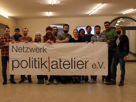 Das Bild zeigt eine Gruppe junger Erwachsener mit Migrationserfahrung in einem Flur der Volkshochschule Bonn, die ein großflächiges Banner mit dem Logo des Vereins halten.