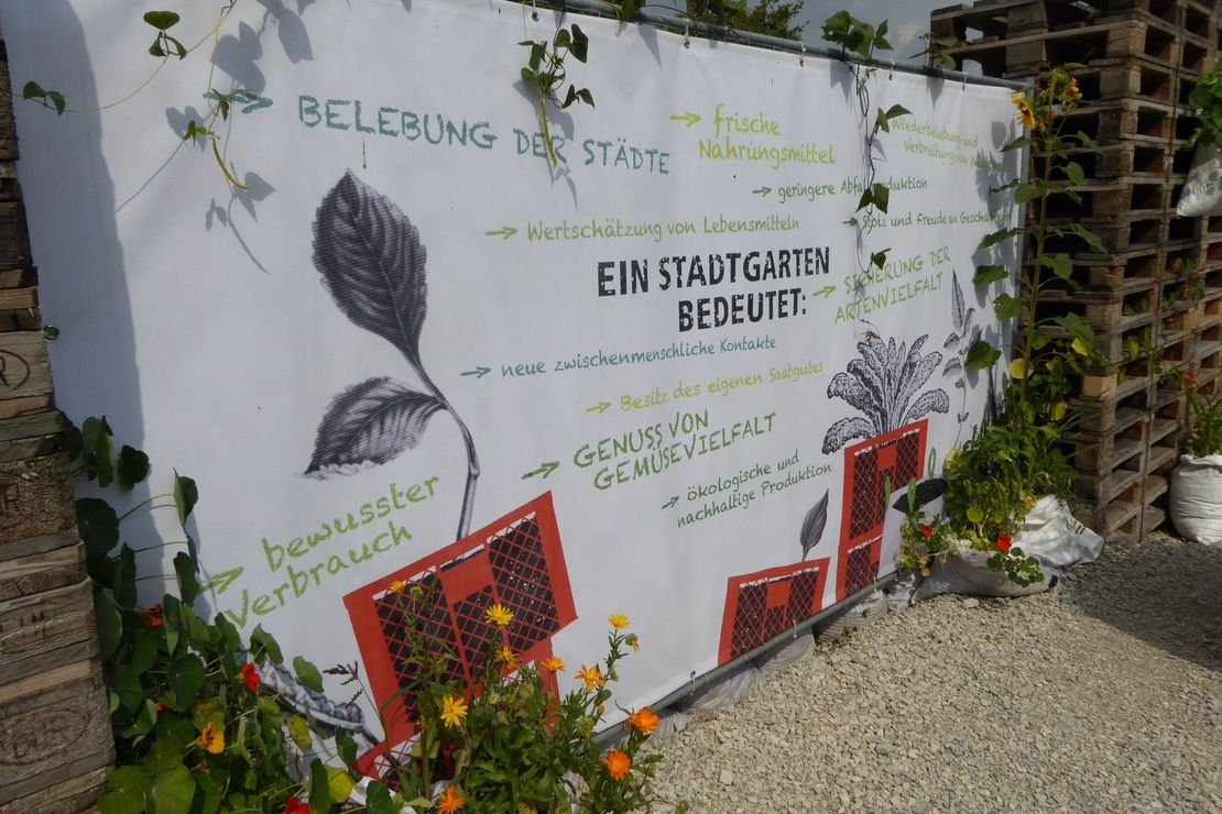 Eine weiße Leinwand zeigt den Titel "Ein Stadtgarten bedeutet". Ebenfalls zu lesen sind unter anderem die Antworten "Genuss von Gemüse", "bewusster Verbrauch", "Wertschätzung von Lebensmitteln" und "neue zischenmenschlich Kontakte". Die Leinwand steht auf einem Schotterboden und ist umringt von Europaletten.
