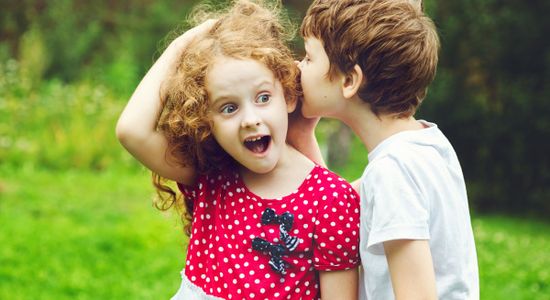 Ein Kind flüstert einem anderen etwas ins Ohr, was überrascht und erfreut reagiert