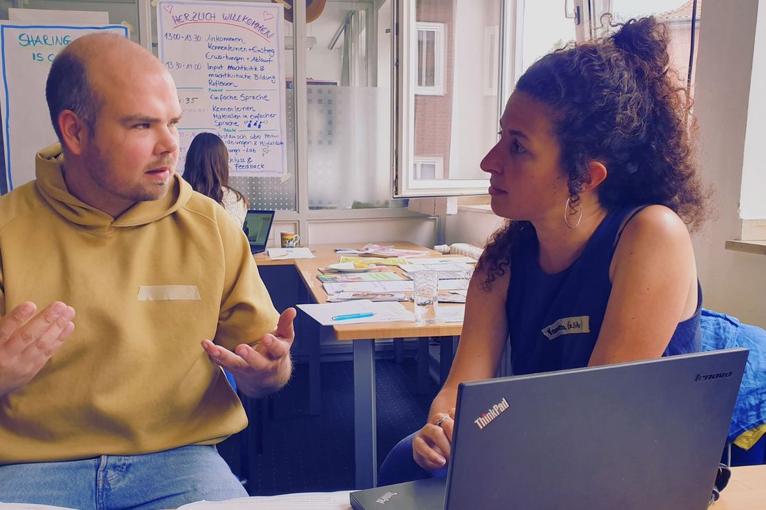 Auf dem Bild sind zwei Personen zu sehen, die im Gespräch sind. Im Vordergrund steht ein Laptop, im Hintergrund sind Plakate und ein Büchertisch zu sehen