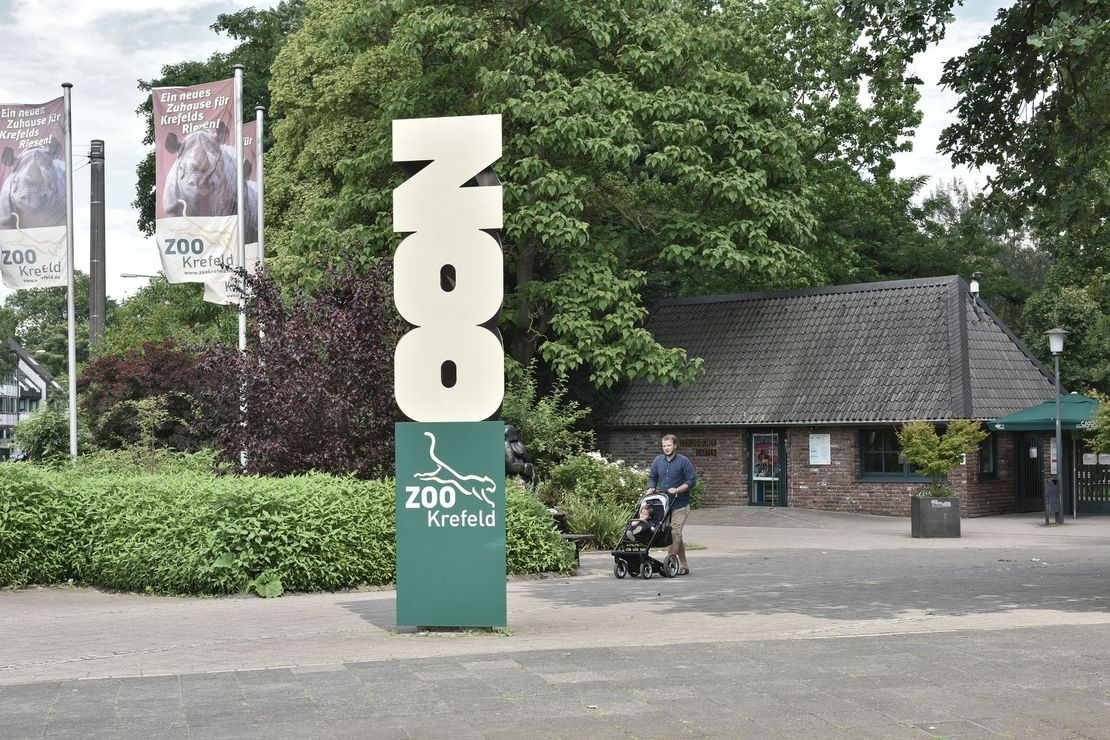 Zu sehen ist der Eingangsbereich des Zoo Krefeld mit Besuchern, Kassenhäuschen und Fahnen.