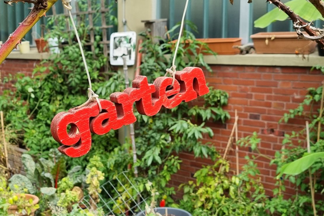 Roter Schriftzug Garten aus Holz aufgehängt vor einer grün berankten Backsteinwand