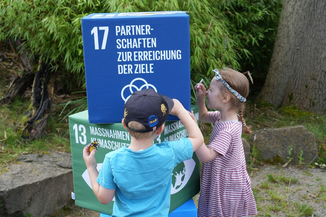 Zwei Kinder untersuchen mit zwei Lupen Sitzwürfel mit den aufgedruckten Bildern von SDG 13 und 17.