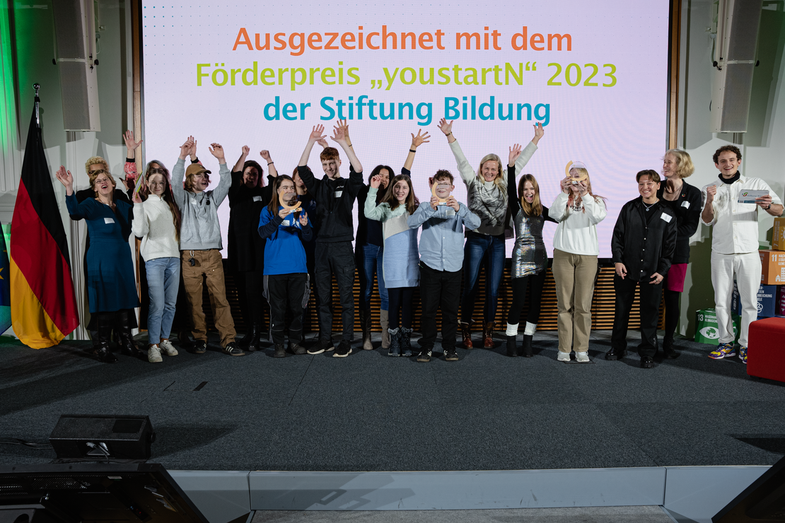 Das Foto zeigt eine Bühne auf der Menschen stehen. Kinder und Jugendliche heben lächelnd ihre Auszeichnungen in die Luft.